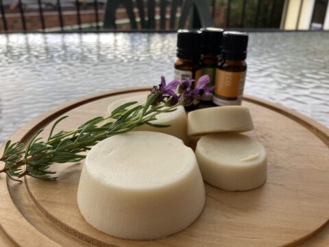 image shows a round wooden board with a small pile of round lotion bars of different sizes, three small essential oil bottles, and a small bunch of lavender