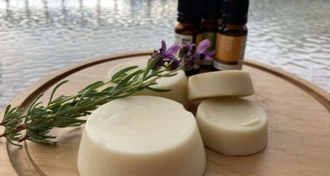 image shows a round wooden board with a small pile of round lotion bars of different sizes, three small essential oil bottles, and a small bunch of lavender